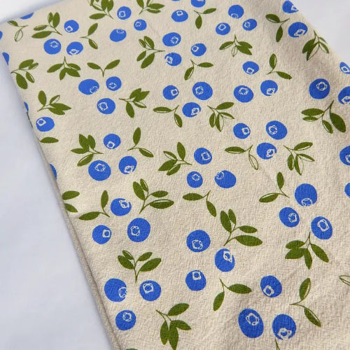 Beige tea towel with blue blueberries and green leaves patterns on an off-white background.