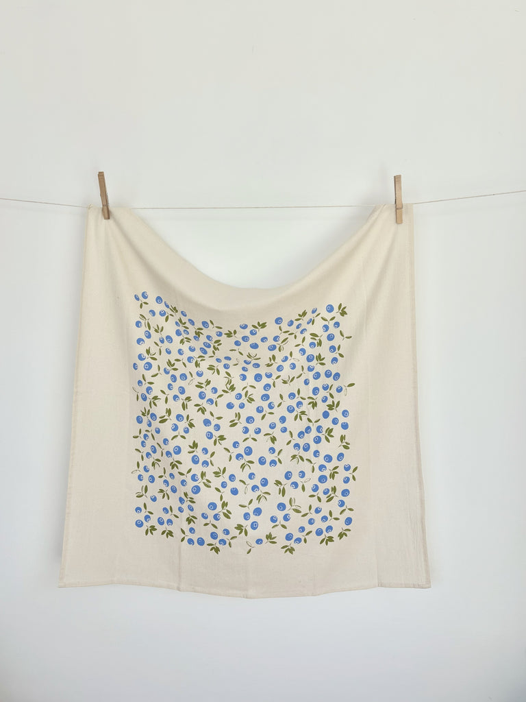 A natural cotton tea towel with blue blueberries and green leaves patterns hanging on a clothesline.