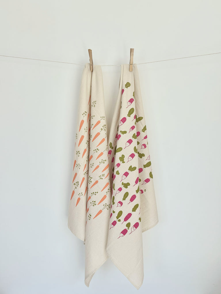 Two natural cotton tea towels with carrot and radish patterns hanging on a clothesline.