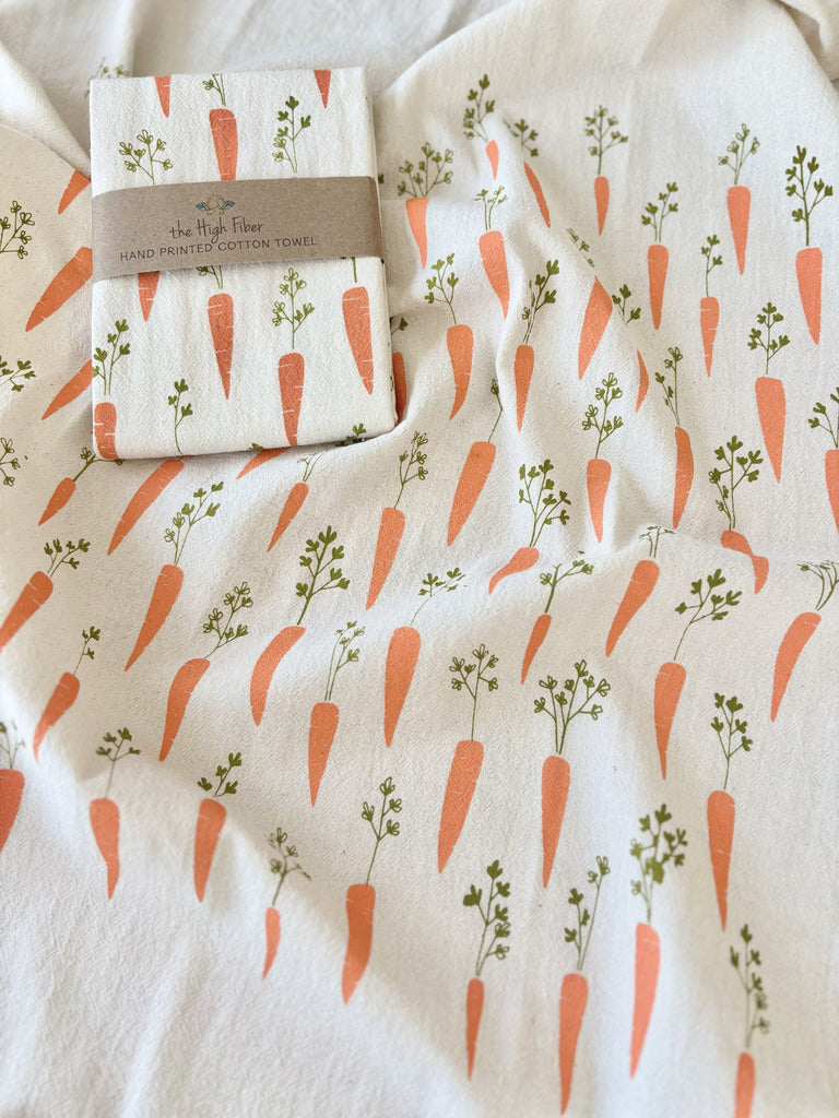 A natural cotton tea towels with orange carrots patterns, with another tea towel wrapped in a brown paper band with "the High Fiber" and the logo printed on it. 