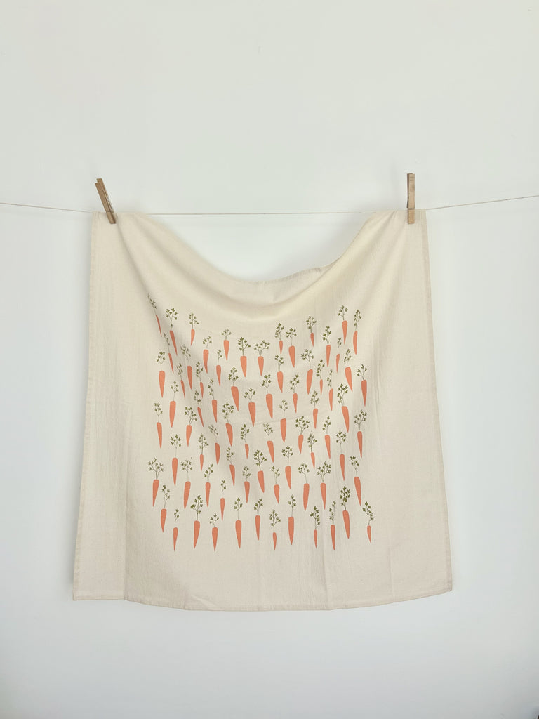 A natural cotton tea towel with orange carrots patterns hanging on a clothesline.  