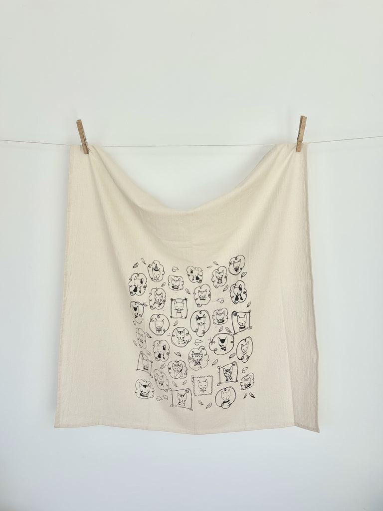 A natural cotton tea towel with various cats portrait patterns hanging on a clothesline. 