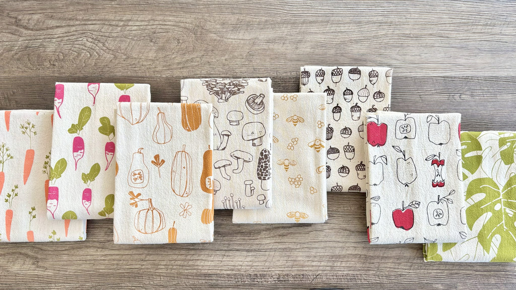 A variety of patterned natural cotton tea towels are displayed on a wooden surface.