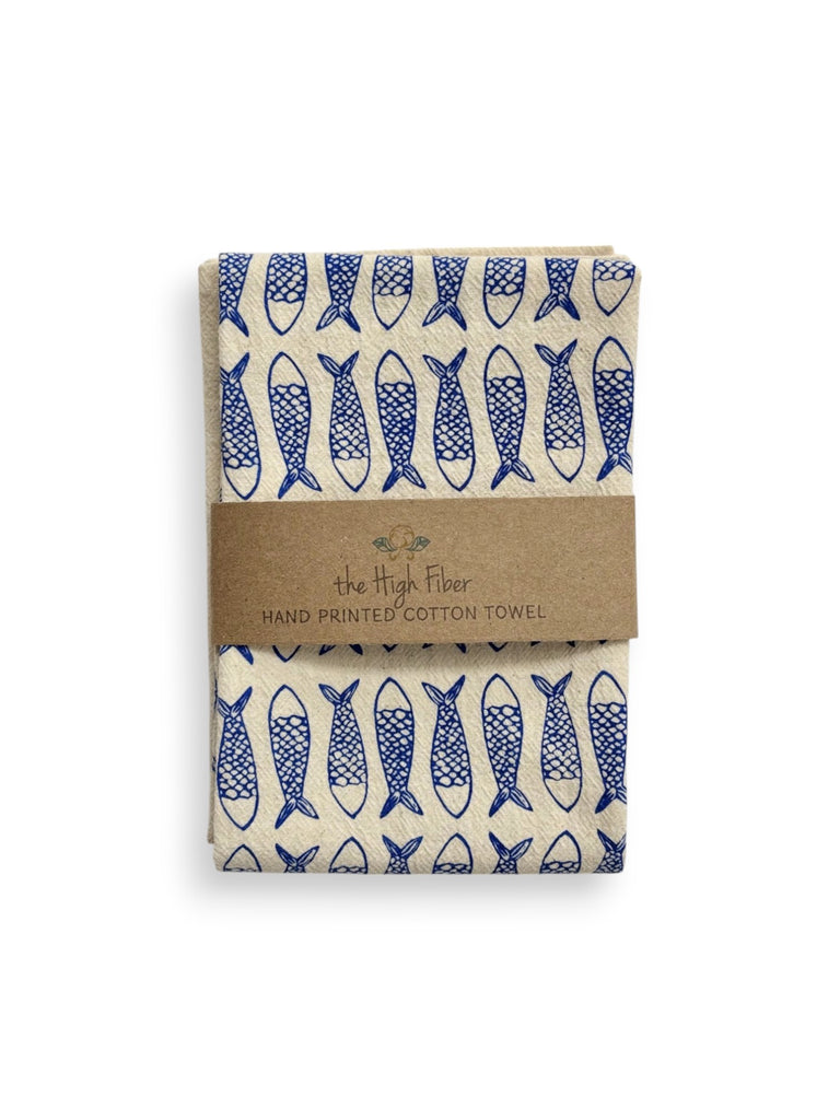 A folded beige tea towel with blue fish patterns and a brown paper band with brand name "the High Fiber" and logo.