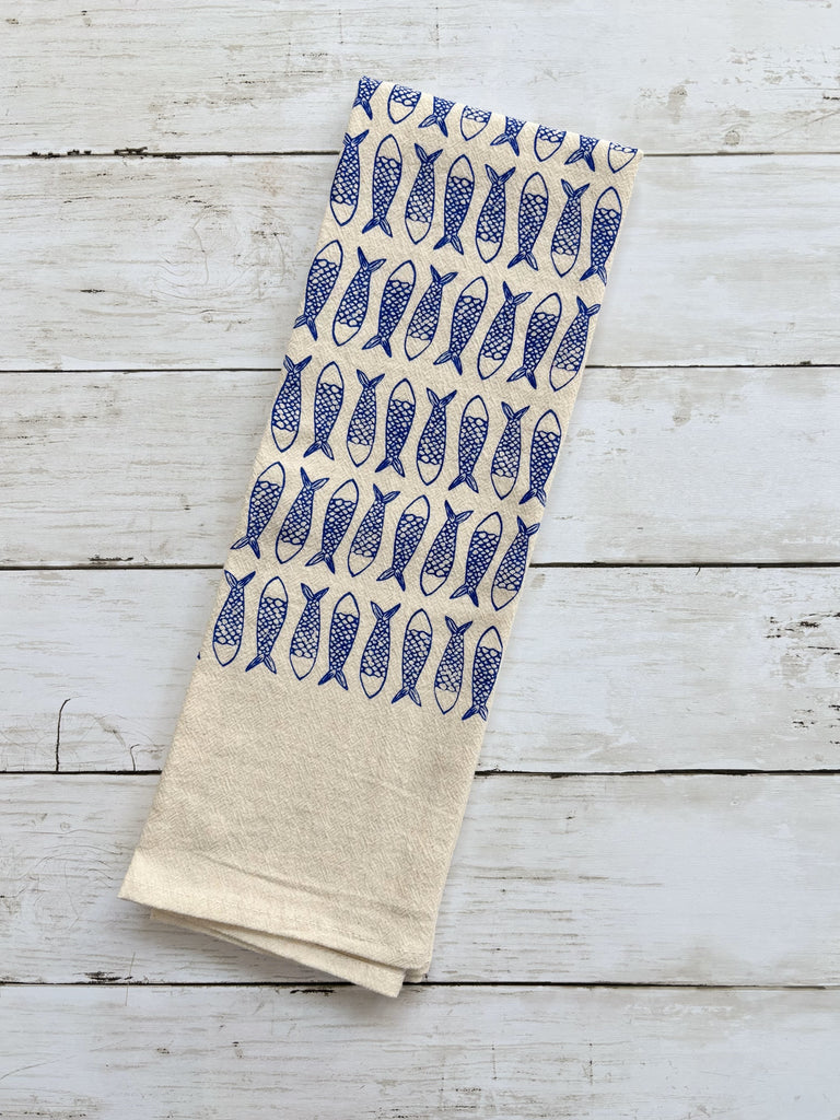 Beige tea towel with blue fish patterns on whitewashed wood surface.