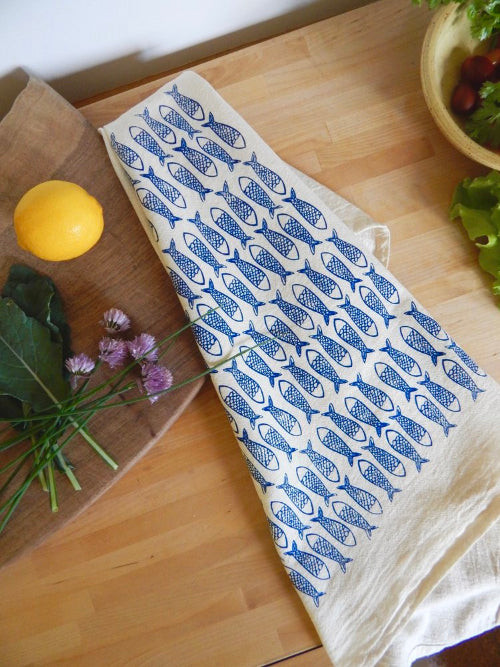 Beige tea towel with blue fish patterns on a wooden countertop with lemon, greens, and flowers.