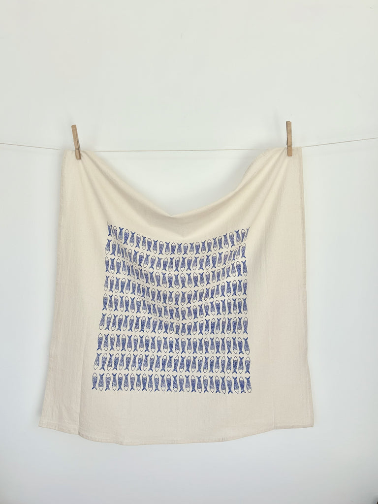 A natural cotton tea towel with blue fishes patterns hanging on a clothesline.
