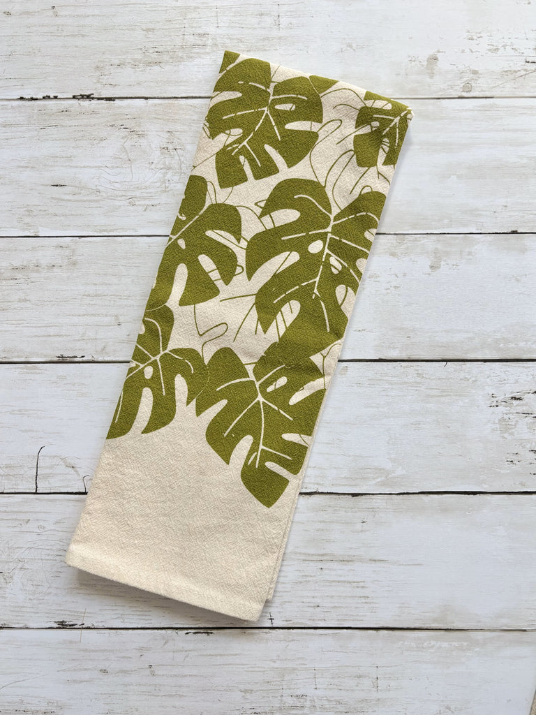 Beige tea towel with green monstera leaves patterns on whitewashed wood surface.