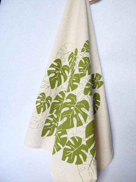 A beige tea towel with green monstera leaves patterns hanging on a white background.