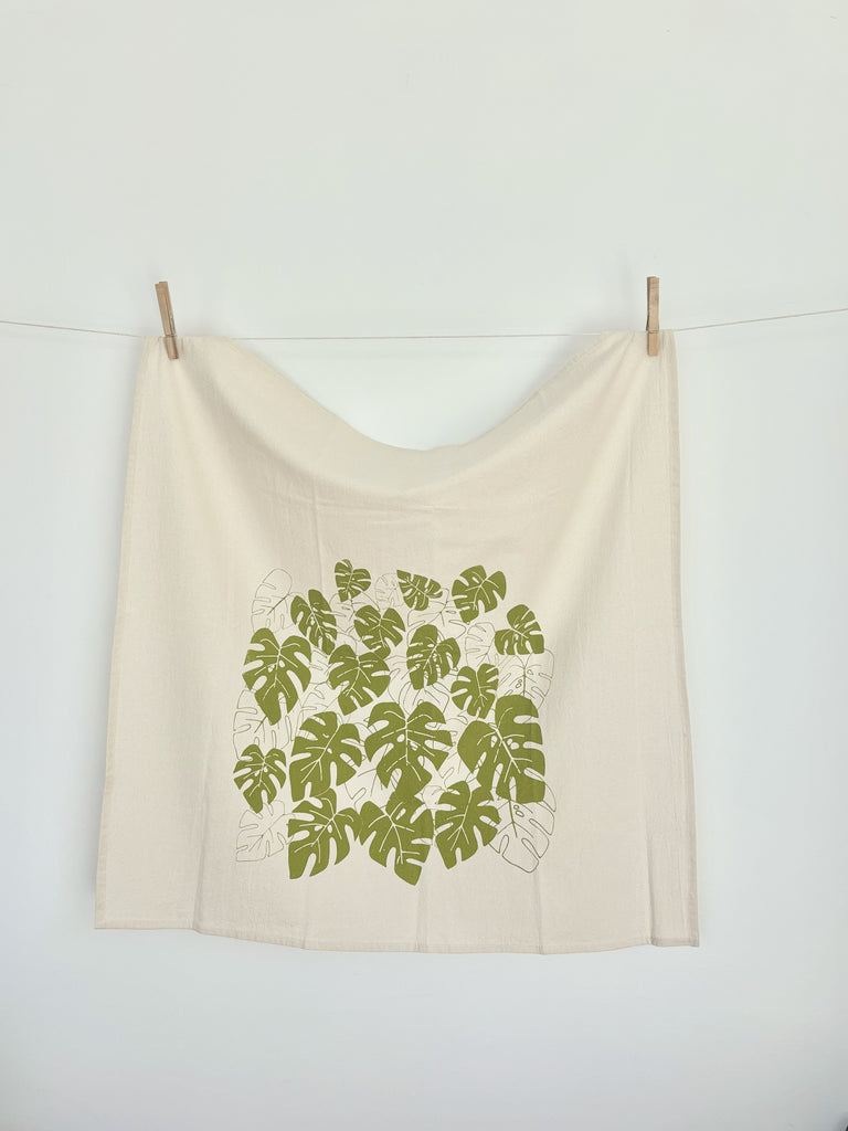 A natural cotton tea towel with green monstera leaves patterns hanging on a clothesline.