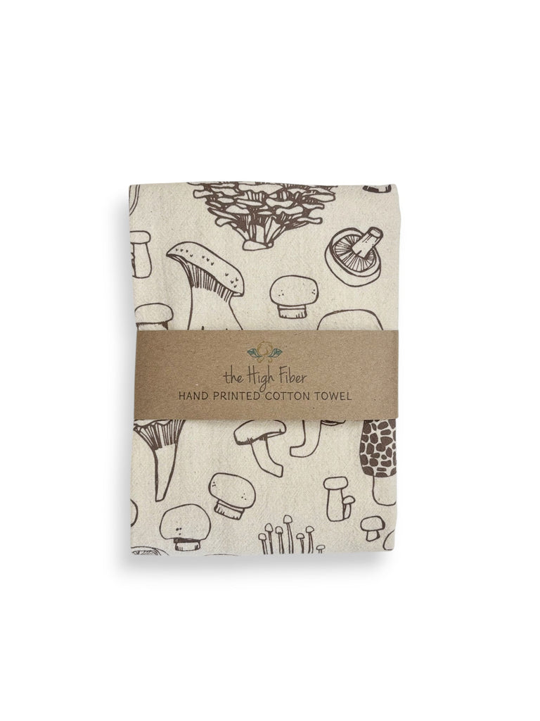 A folded natural cotton tea towel with taupe mushrooms patterns and a brown paper band with brand name "the High Fiber" and logo.