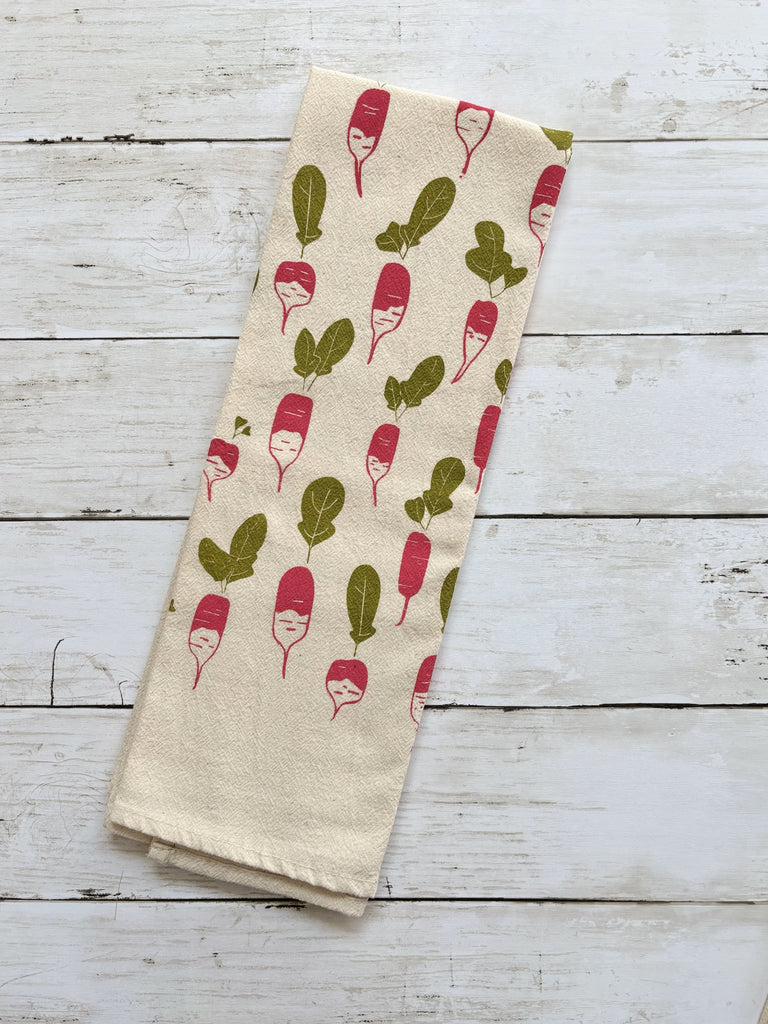 Beige tea towel with red radishes and green leaves patterns on whitewashed wood surface.