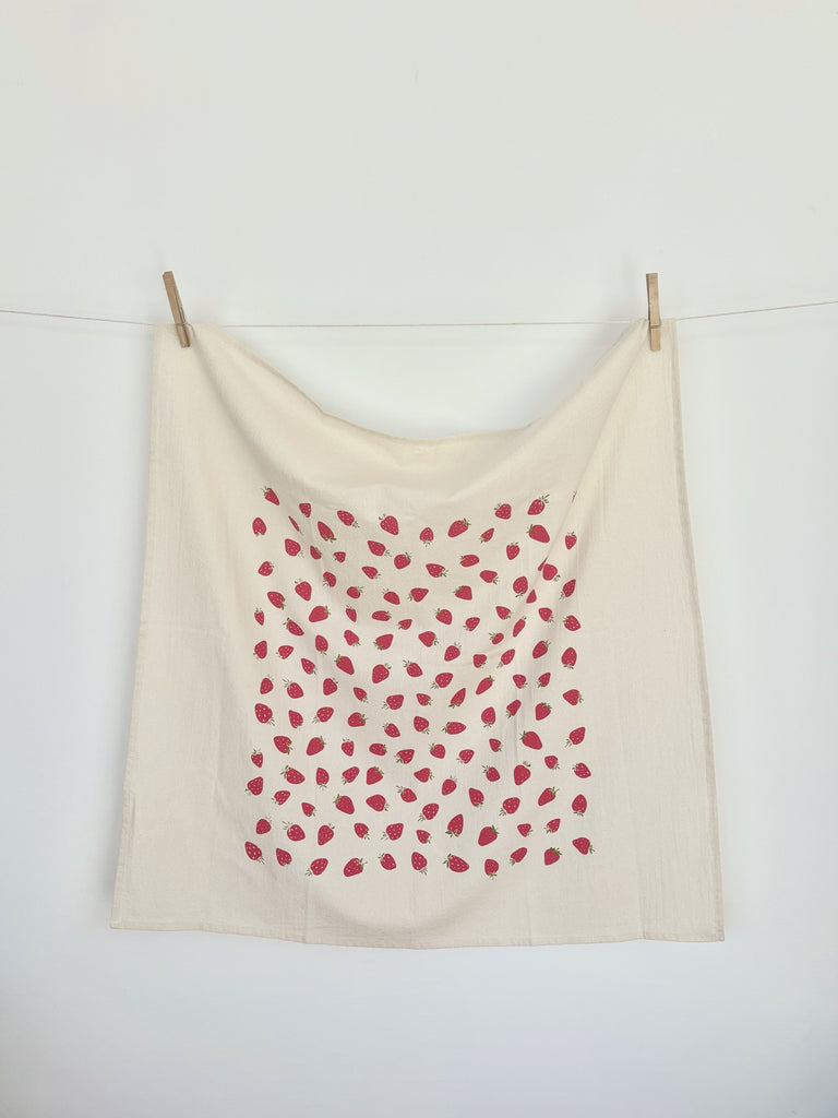 A natural cotton tea towel with red strawberries patterns hanging on a clothesline.