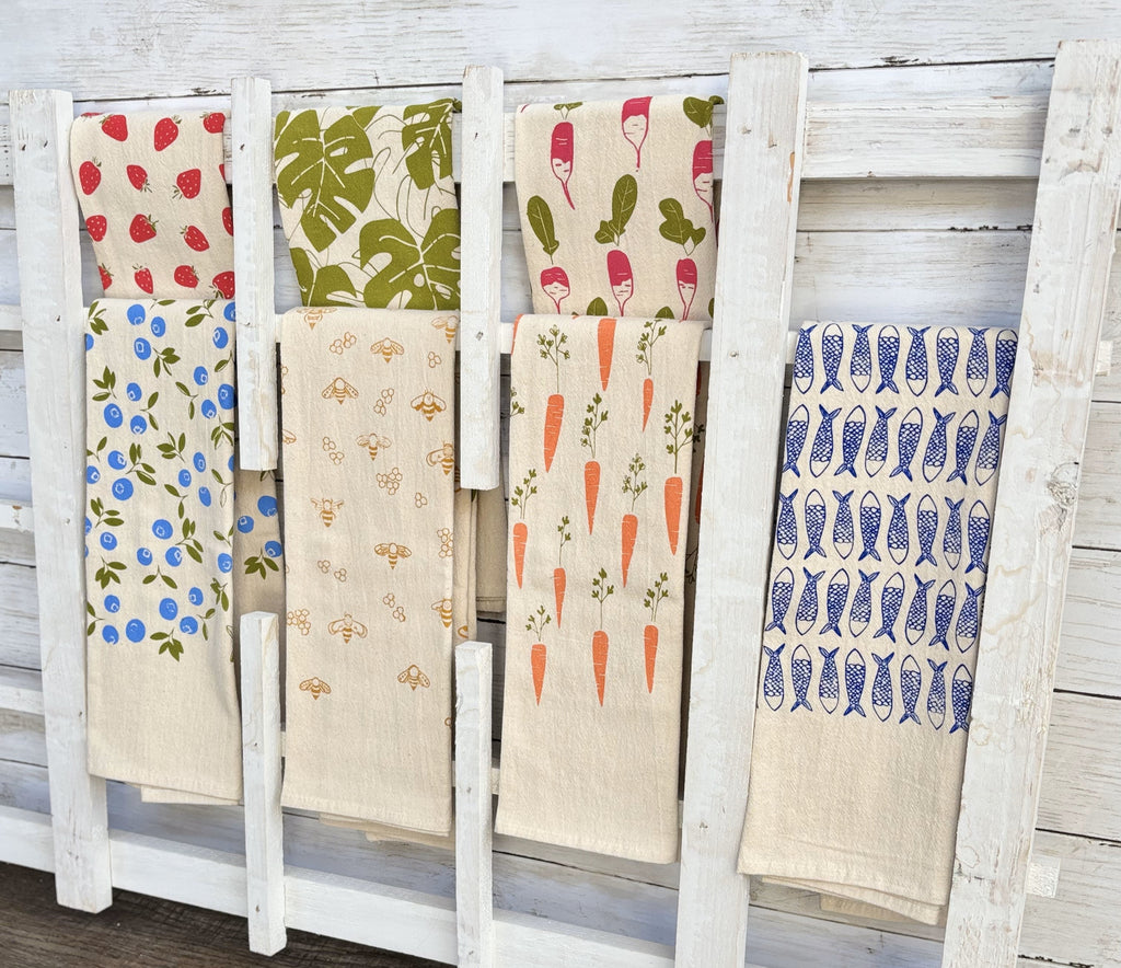 A rack displaying assorted hand-printed tea towels with designs of strawberries, blueberries, monstera leaves, radishes, bees, carrots, and fish patterns, against a white wooden wall.