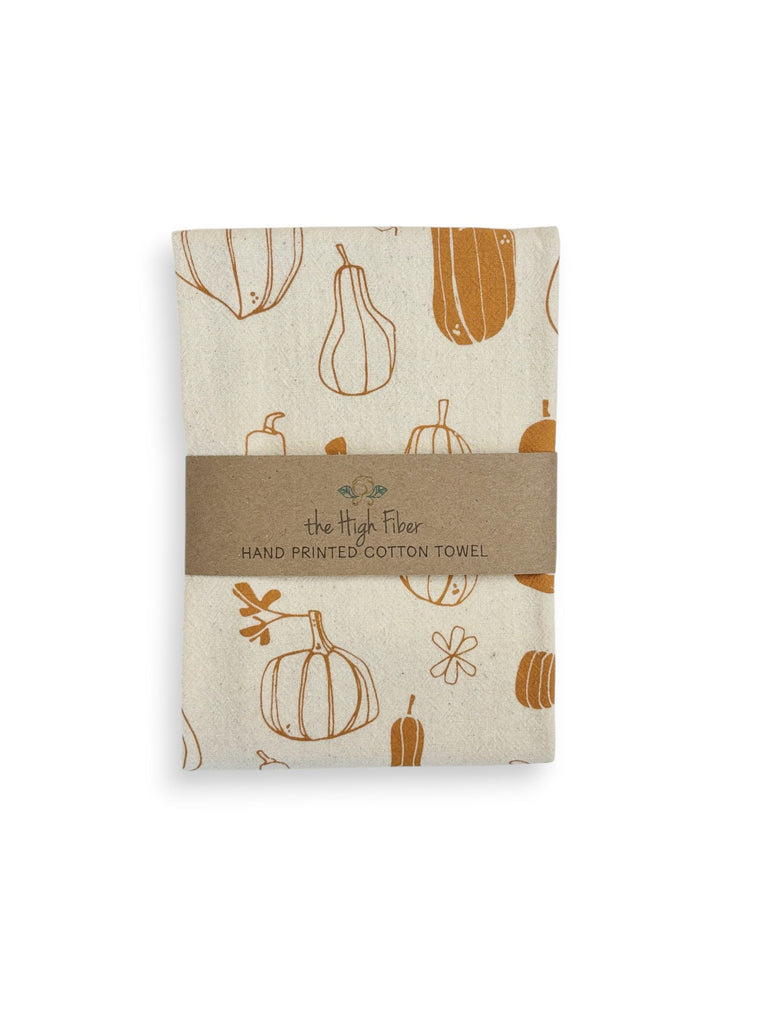 A folded natural cotton tea towel with burnt orange winter squashes and pumpkins patterns and a brown paper band with brand name "the High Fiber" and logo.