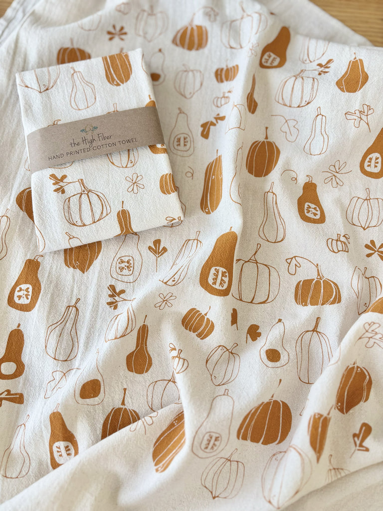 A natural cotton tea towels with burnt orange winter squashes and pumpkins patterns, with another tea towel wrapped in a brown paper band with "the High Fiber" and the logo printed on it. 