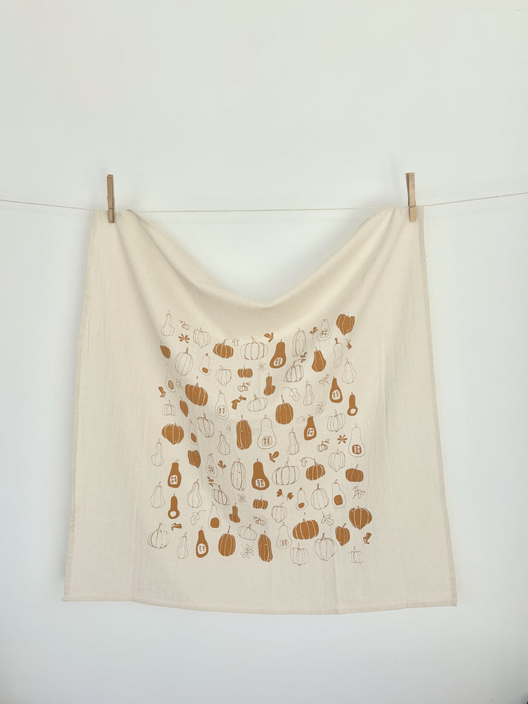 A natural cotton tea towel with burnt orange winter squashes and pumpkins patterns hanging on a clothesline.
