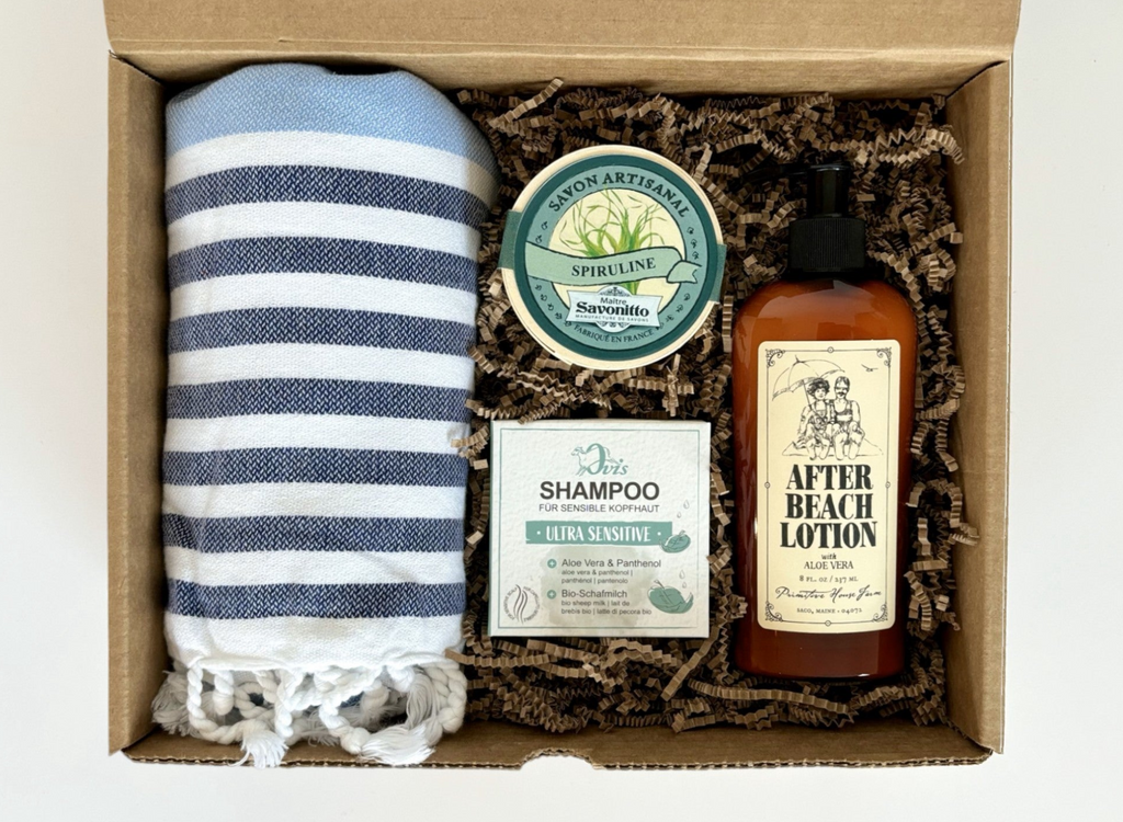 Top view of a gift box containing a striped towel, a round soap in wood box, a shampoo in box, and a brown bottle of lotion.