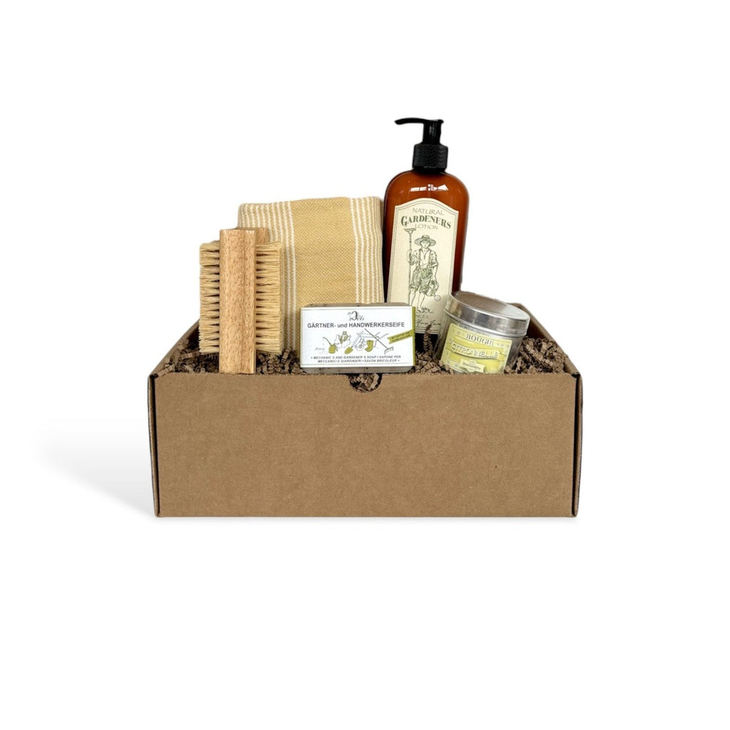 Gift box with personal care for gardeners, including a nail brush, a striped towel, a soap bar, a brown bottle of lotion, and a candle tin.