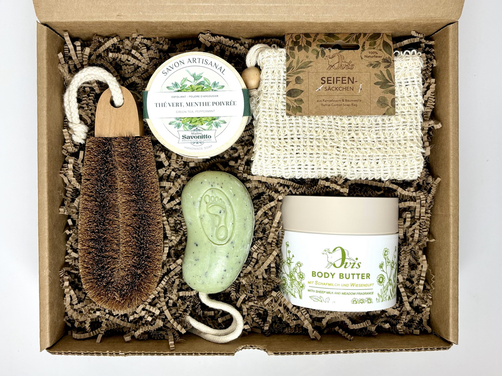 Top view of a gift box of spa products including a foot scrub soap bar, a round soap in wood box, a body butter, a soap bag and a foot brush.
